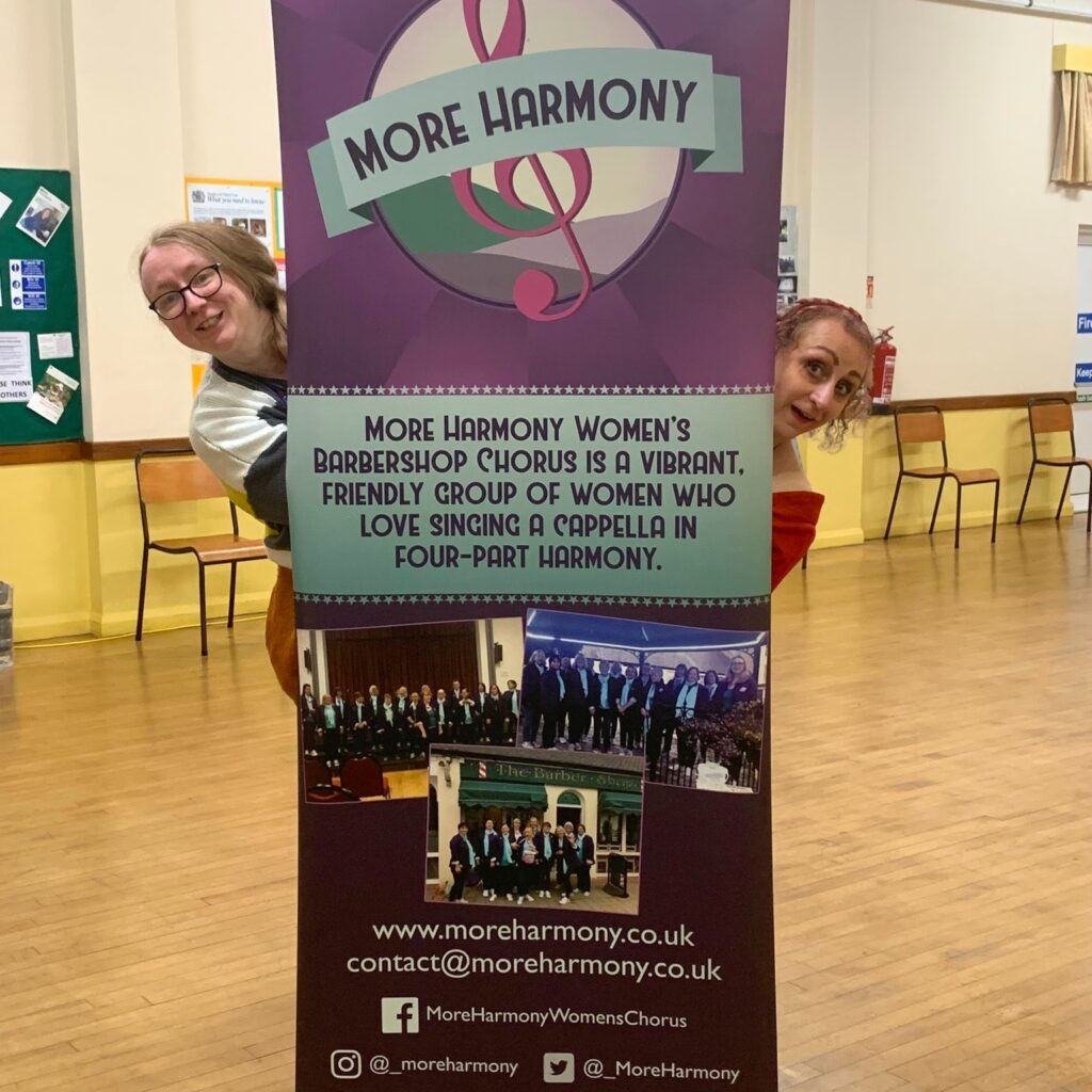 Emma & Lesley with a banner for More Harmony Women's Barbershop Chorus, designed by Little Bird Creative